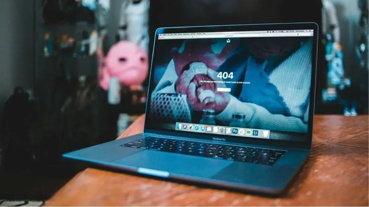 Ordinateur portable MacBook posé sur une table en bois affichant une page d'erreur 404 (page introuvable), suggérant un problème de site, avec l'arrière-plan flou montrant une peluche rose et un coin de bureau.