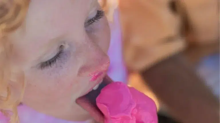 Jeune personne rousse dégustant une grosse glace rose sur un cornet, le rose fondant couvrant ses lèvres et ses mains, expression concentrée et ambiance estivale détendue.