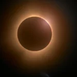 Éclipse solaire totale montrant la Lune comme disque sombre masquant le Soleil, entourée d'une couronne solaire orange-dorée et d'un éclat lumineux ponctuel sur la bordure.