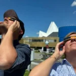 Deux personnes regardant le ciel avec des lunettes solaires d'observation pendant une éclipse, debout sur une pelouse devant des bâtiments modernes (tour bleue et structure blanche angulaire) à la cité de l'espace à Toulousse par une journée ensoleillée.