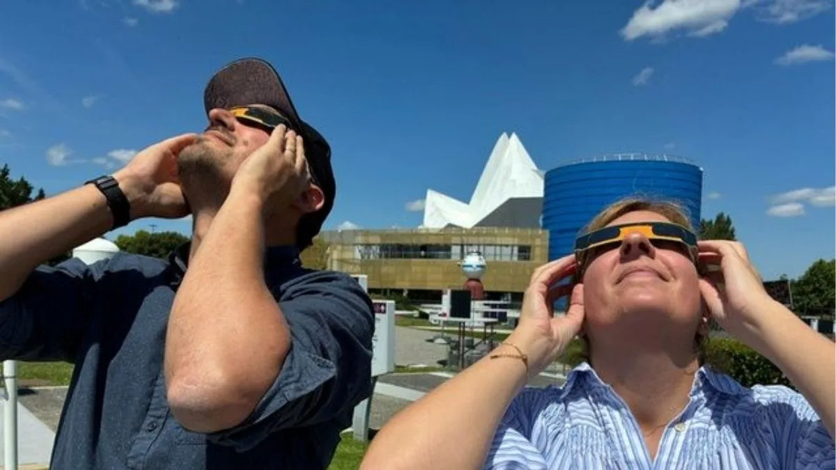 Deux personnes regardant le ciel avec des lunettes solaires d'observation pendant une éclipse, debout sur une pelouse devant des bâtiments modernes (tour bleue et structure blanche angulaire) à la cité de l'espace à Toulousse par une journée ensoleillée.