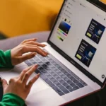 Personne utilisant un ordinateur portable pour concevoir des visuels publicitaires (maquettes d'une chaise) dans un éditeur graphique, mains tapant au clavier assise sur un canapé.