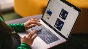 Personne utilisant un ordinateur portable pour concevoir des visuels publicitaires (maquettes d'une chaise) dans un éditeur graphique, mains tapant au clavier assise sur un canapé.