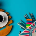 Trousse à crayons à motif d’ancre ouverte sur fond bleu, crayons et onglets colorés qui débordent, à côté d’une tasse de café sur un dessous de bois et d’un petit cactus.
