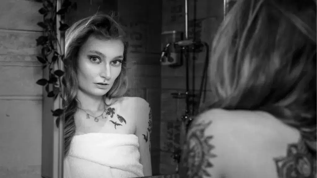 Femme enveloppée d'une serviette regardant son reflet dans un miroir de salle de bains en noir et blanc, montrant des tatouages sur l'épaule et la clavicule, avec robinetterie et douche en arrière-plan. Photo © Glintof.