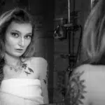 Femme enveloppée d'une serviette regardant son reflet dans un miroir de salle de bains en noir et blanc, montrant des tatouages sur l'épaule et la clavicule, avec robinetterie et douche en arrière-plan. Photo © Glintof.