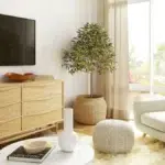 Coin salon lumineux et chaleureux conçu pour la détente : fauteuil beige confortable, pouf tricoté, commode en bois clair, grande plante en pot, table basse avec vase et livre, télé murale et baie vitrée laissant entrer la lumière naturelle.