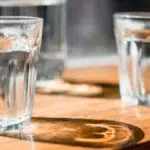 Trois verres d'eau posés sur une table en bois, éclairés par le soleil qui traverse le verre et projette des reflets lumineux et des ombres circulaires.