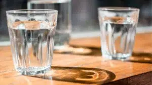 Trois verres d'eau posés sur une table en bois, éclairés par le soleil qui traverse le verre et projette des reflets lumineux et des ombres circulaires.