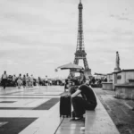 Un voyageur assis, la tête posée sur ses bras près d'une valise au Trocadéro, face à la foule et à la tour Eiffel en arrière-plan, scène en noir et blanc évoquant solitude et fatigue.