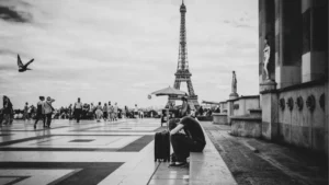 Un voyageur assis, la tête posée sur ses bras près d'une valise au Trocadéro, face à la foule et à la tour Eiffel en arrière-plan, scène en noir et blanc évoquant solitude et fatigue.